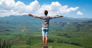 A man stands triumphantly with arms open, overlooking a scenic mountain range.